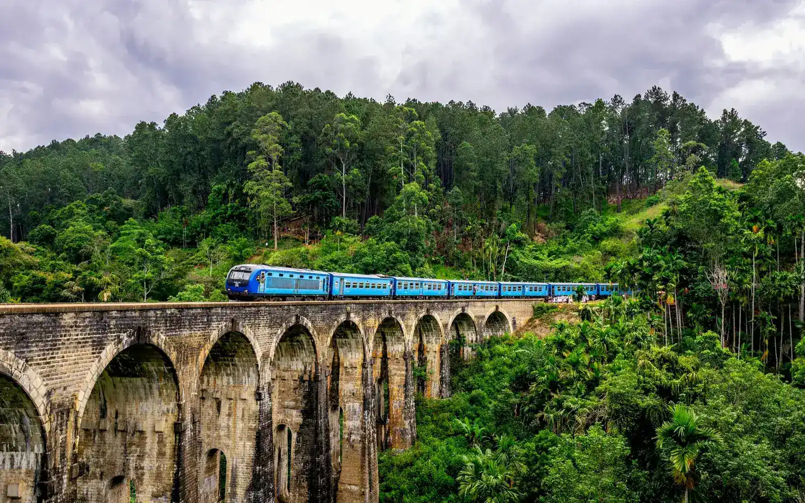 A blue train crossing a stone viaduct through green hills